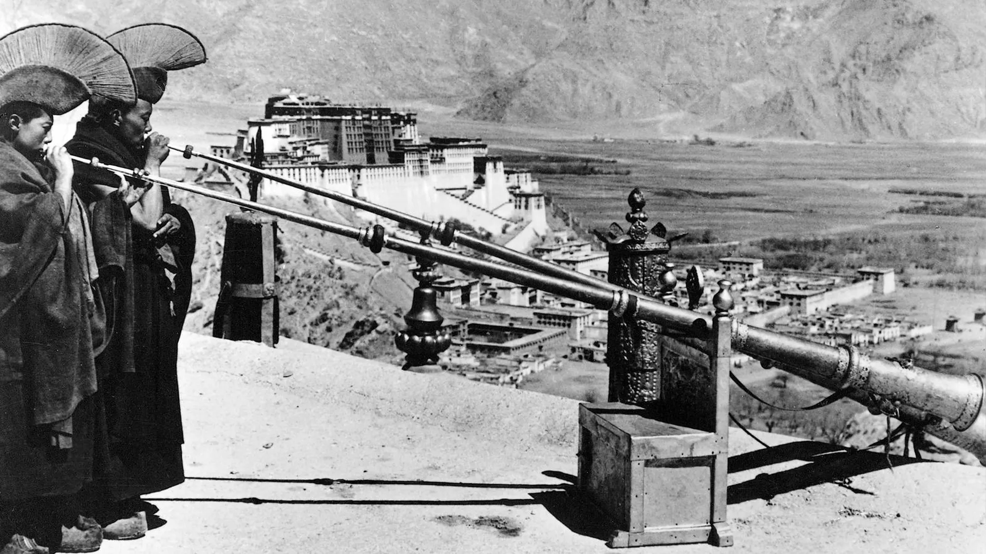 Двое буддийских монахов трубят в рог, на заднем плане — дворец Потала, который был резиденцией Далай-ламы до 1959 года. Кадр из документального немецкого фильма «Тайный Тибет», 1939 год